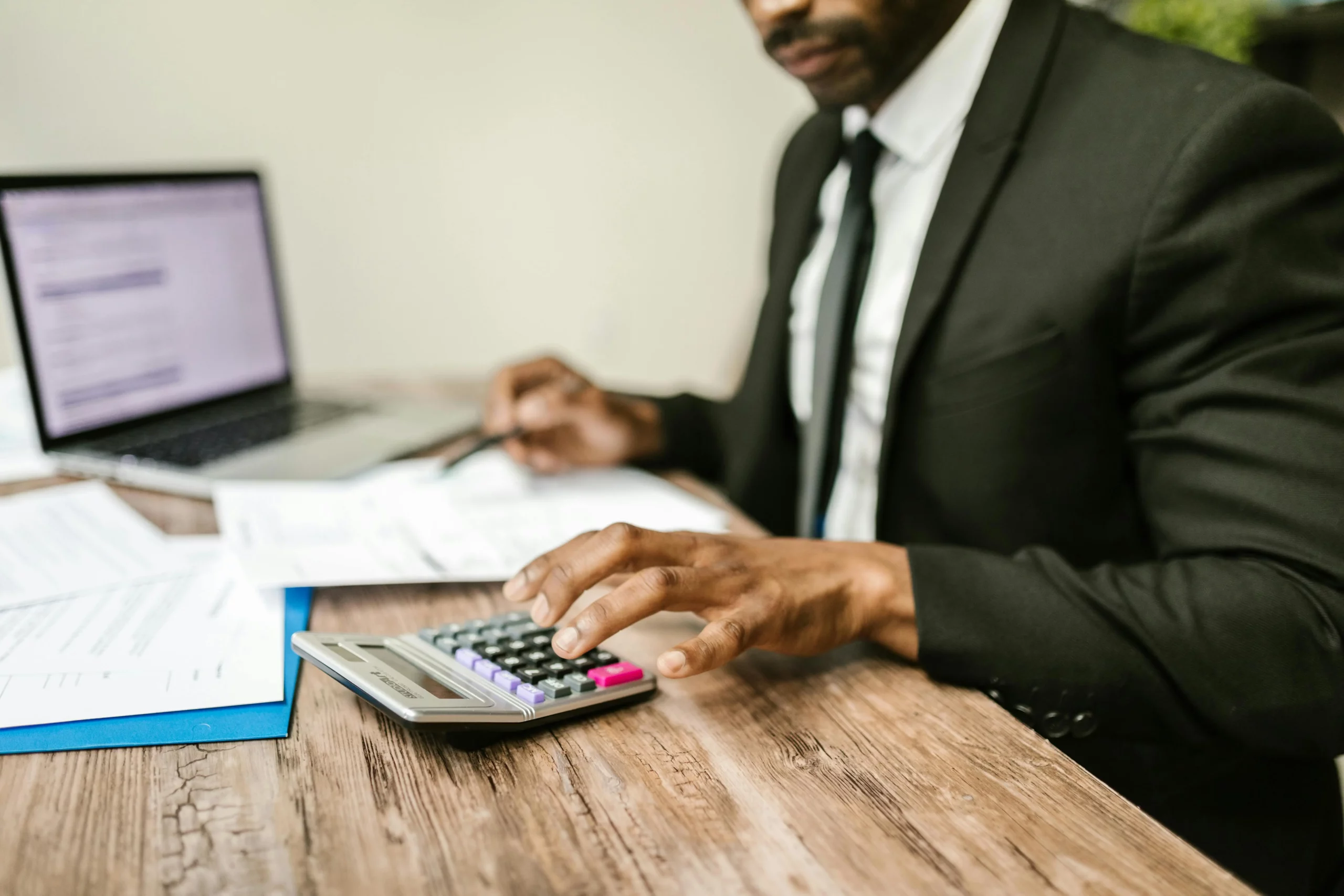 Un homme en costume calcule des finances sur une calculatrice avec des documents et un ordinateur.